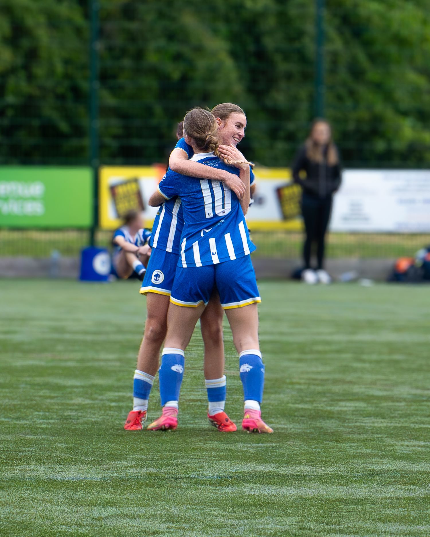 Two female soccer players in blue and white uniforms sharing a celebratory hug on a turf pitch.