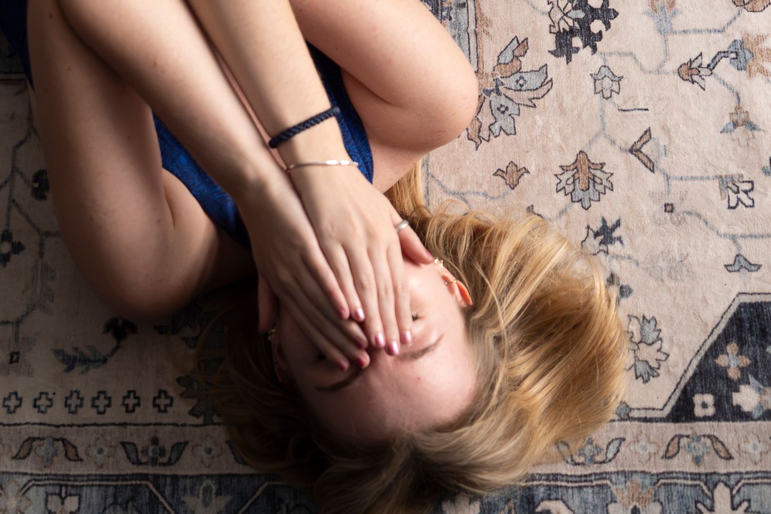 Blonde woman lying on a patterned rug, covering her face with both hands, wearing a blue top.
