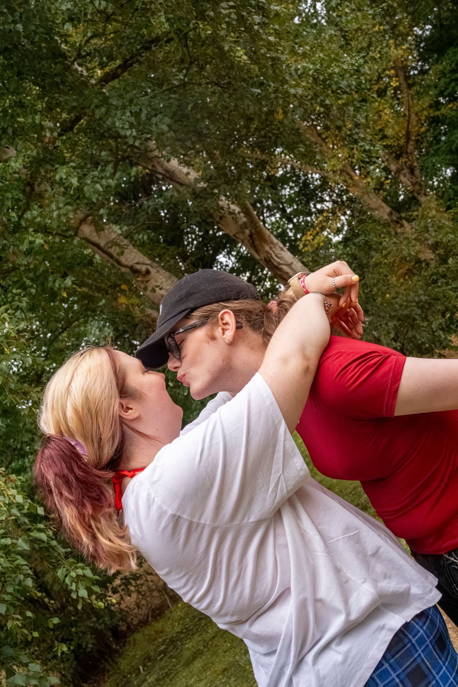Two women sharing a romantic kiss outdoors in a lush green park setting, one wearing a white shirt and black cap.