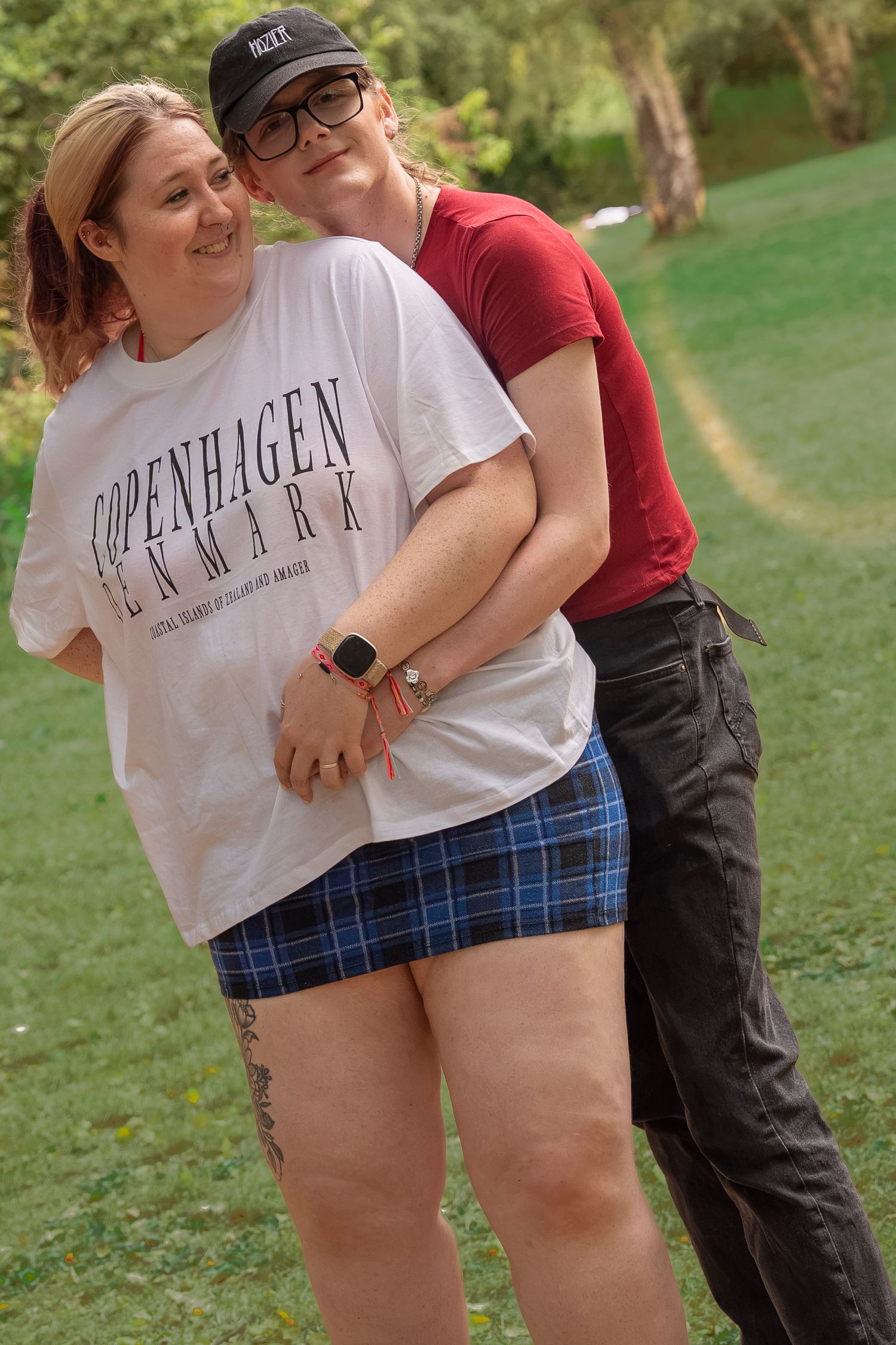 Two women posing outdoors, one wearing a Copenhagen Denmark shirt and plaid skirt, the other in a red top and black pants.