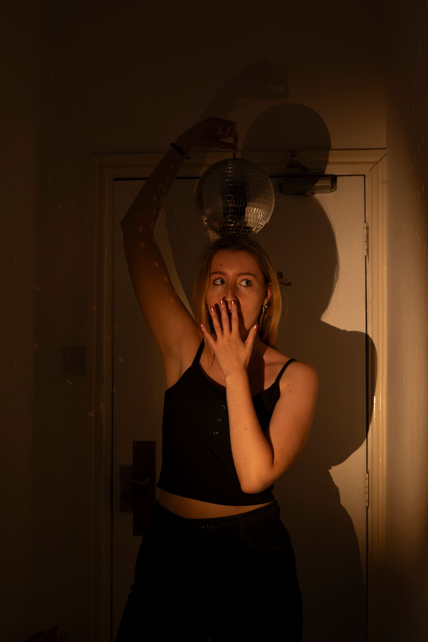 Woman in black crop top holds fan overhead with hand over mouth in dramatic warm golden light near a door.