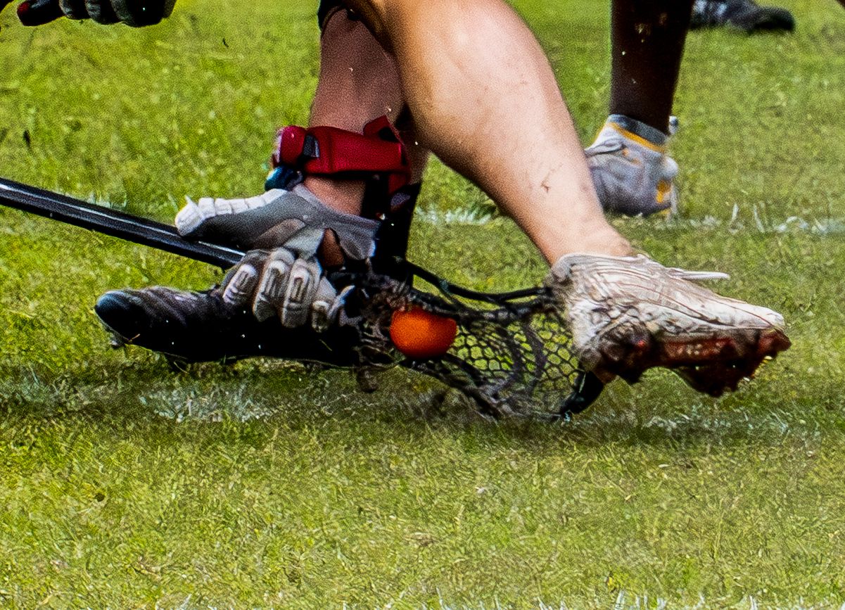 Close-up of lacrosse player's muddy cleats and stick scooping orange ball on grass.