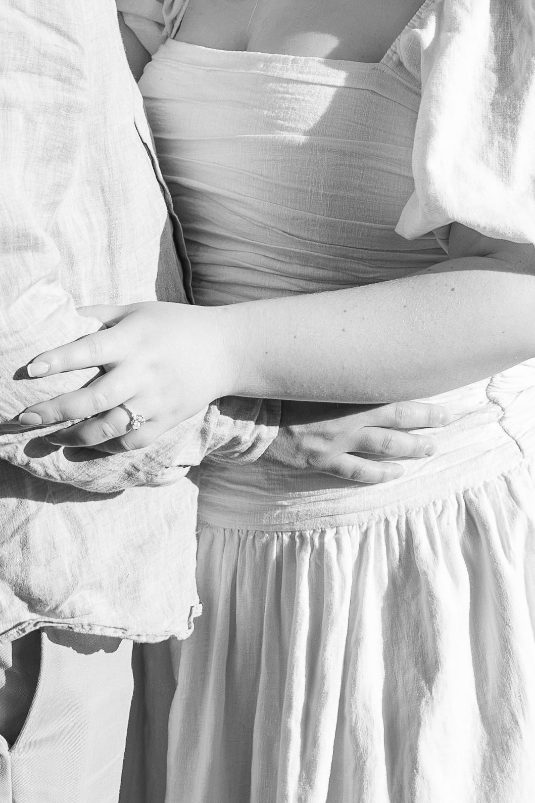 Black and white photo of a couple embracing, showing their hands and light linen clothing in soft natural light.