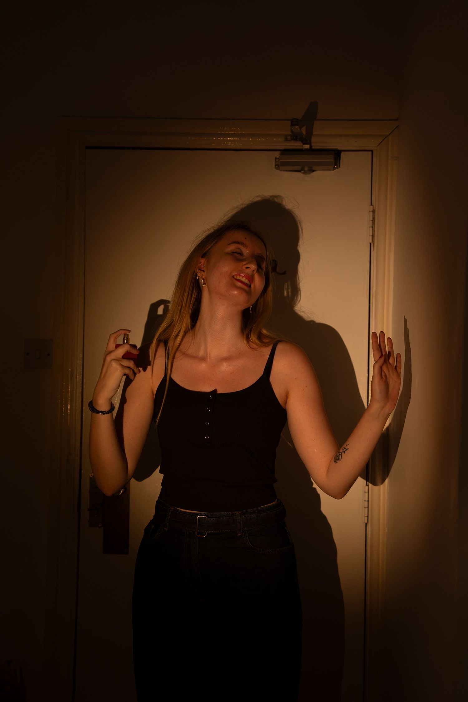 Woman in black tank top posing confidently in warm amber light near a door, holding a phone.
