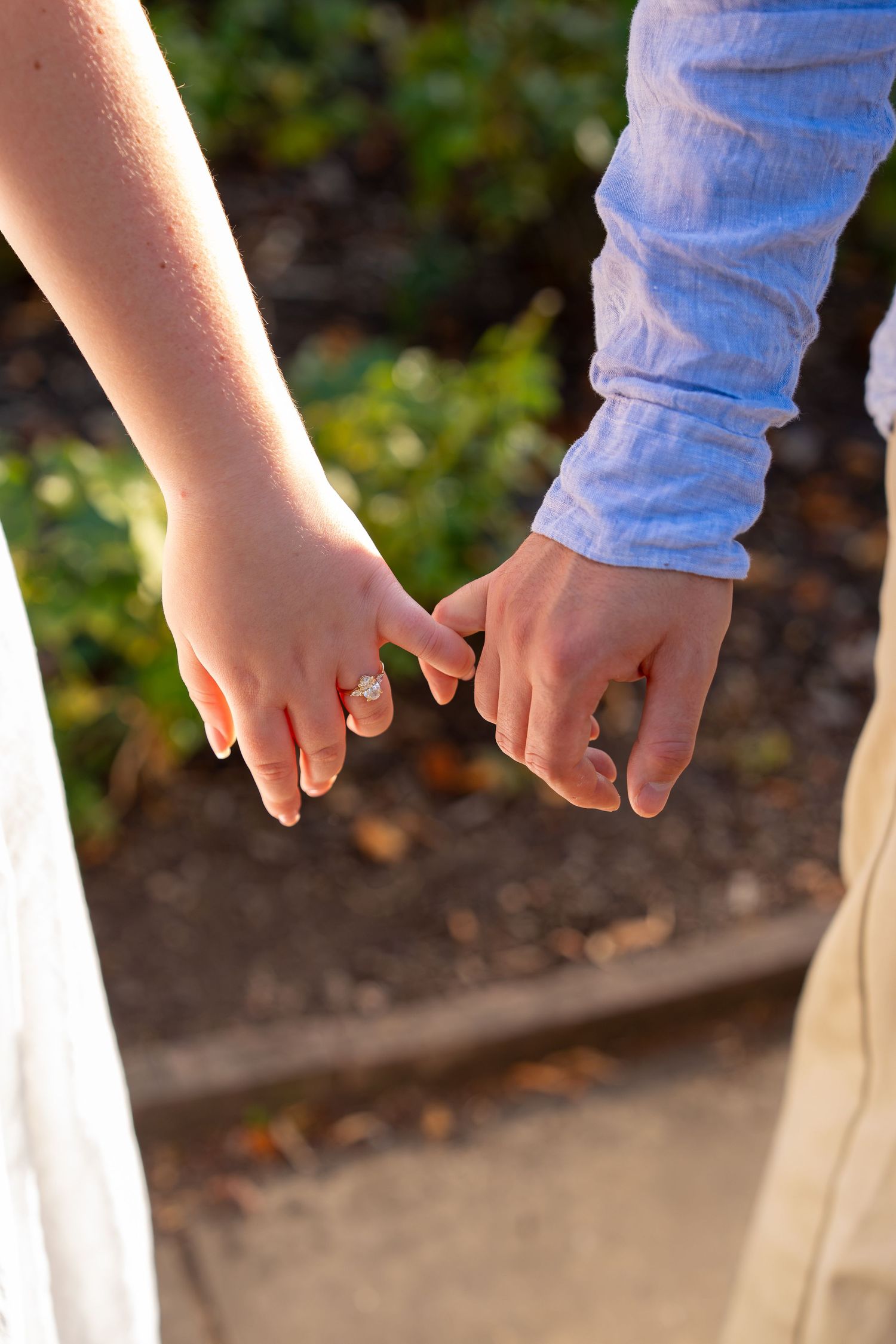 Couple holding hands with engagement ring visible, wearing white dress and blue shirt outdoors.