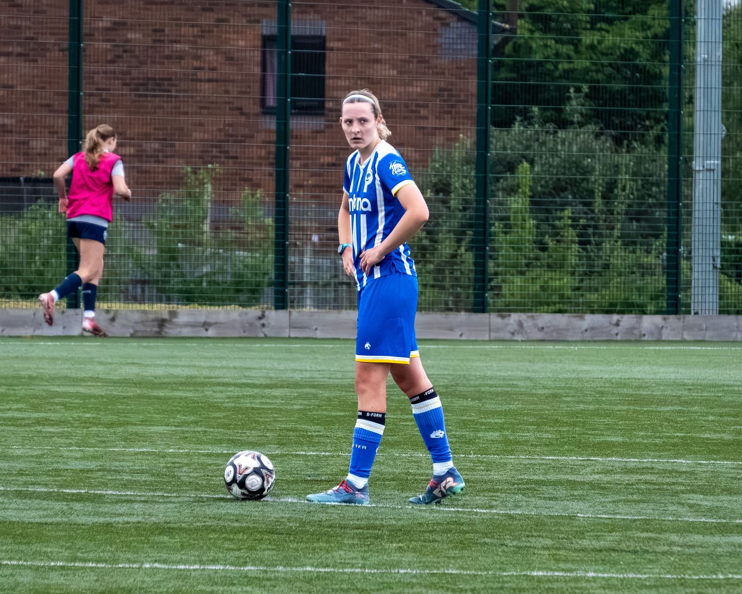 Female soccer player in blue uniform standing with a ball on an artificial turf football pitch.