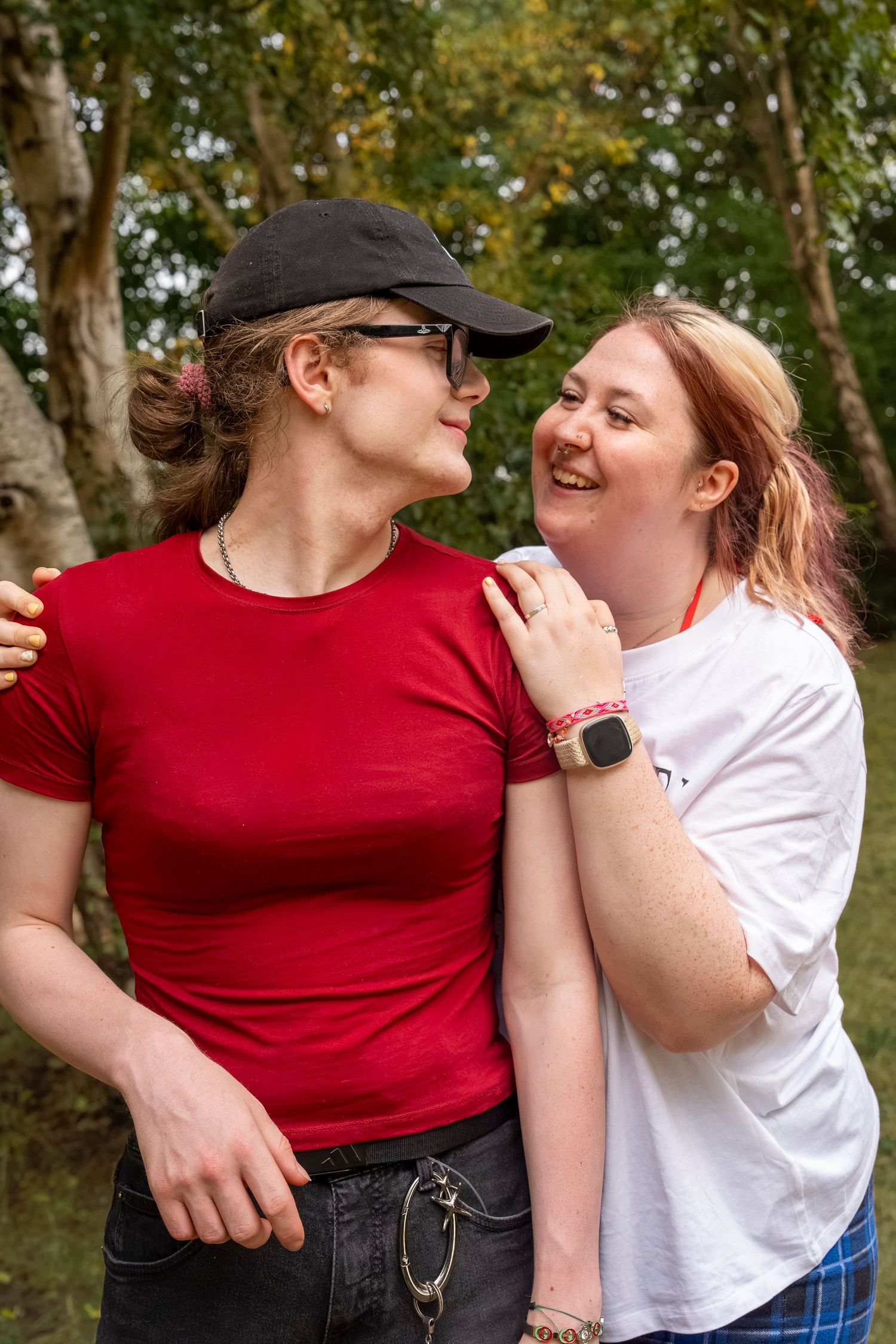 Two young women smiling and embracing outdoors in a park, one wearing a red t-shirt and black cap.