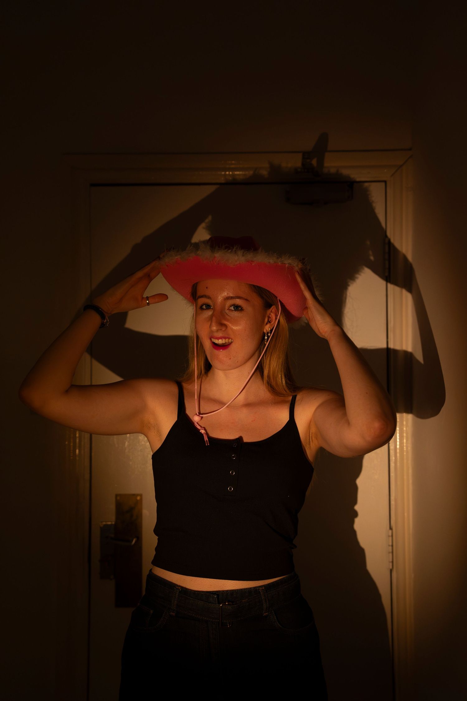 A young woman in a black tank top holds a pink cowboy hat over her head, smiling in warm dim lighting.