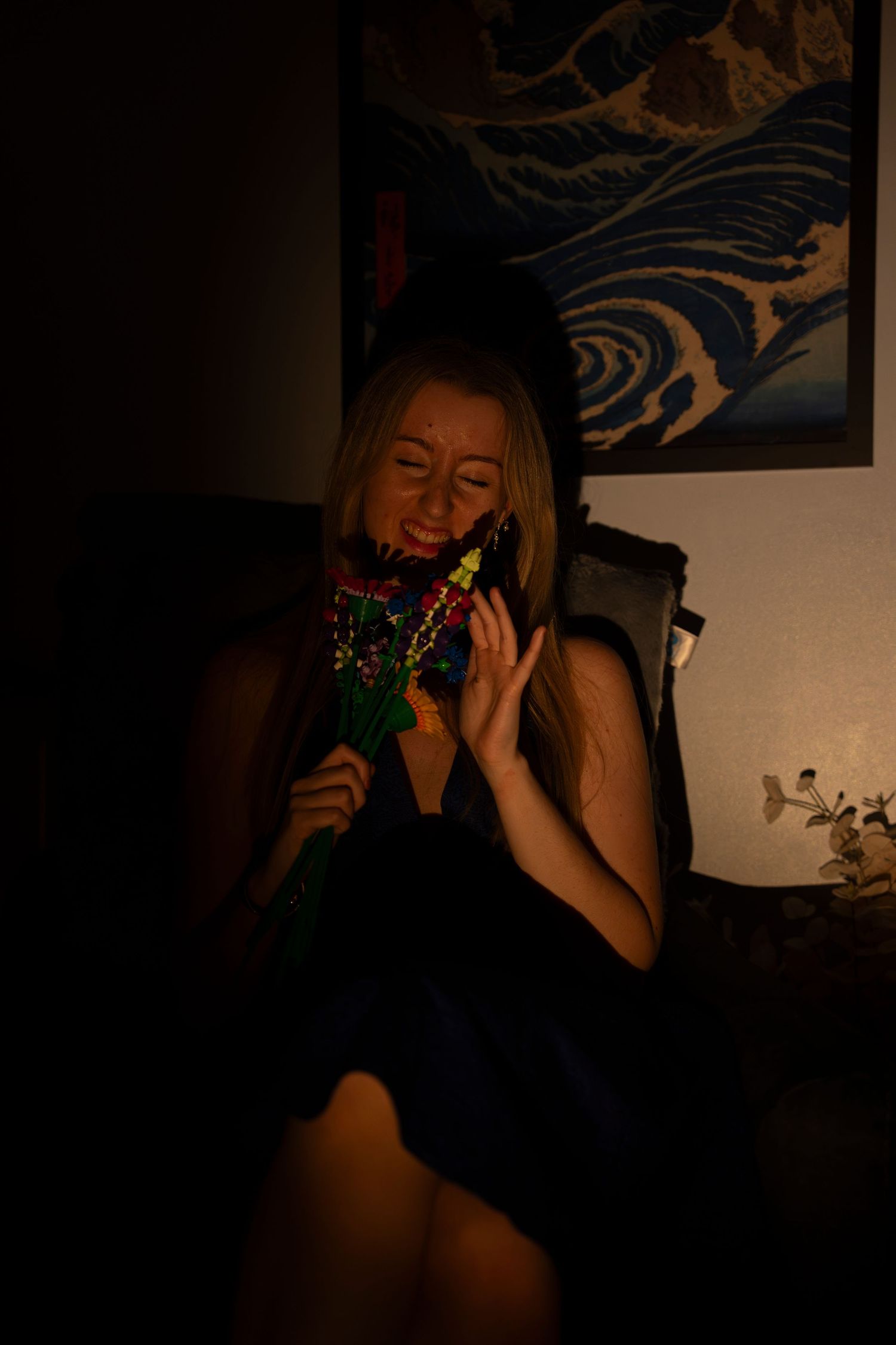 A young blonde woman in a black dress laughs while holding colorful lego flowers in a dimly lit room.