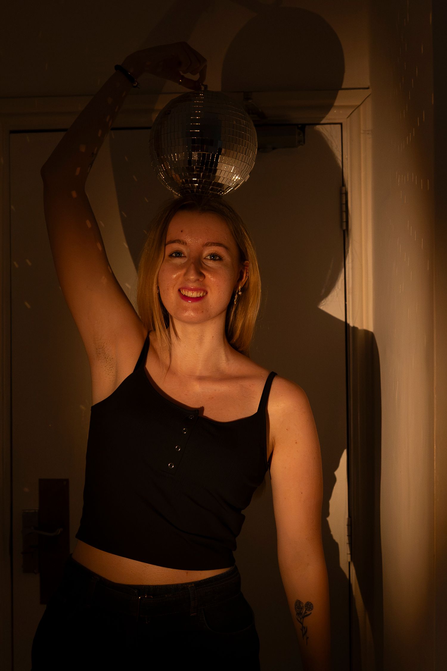 Woman in black crop top holding a disco ball above her head in warm moody lighting.