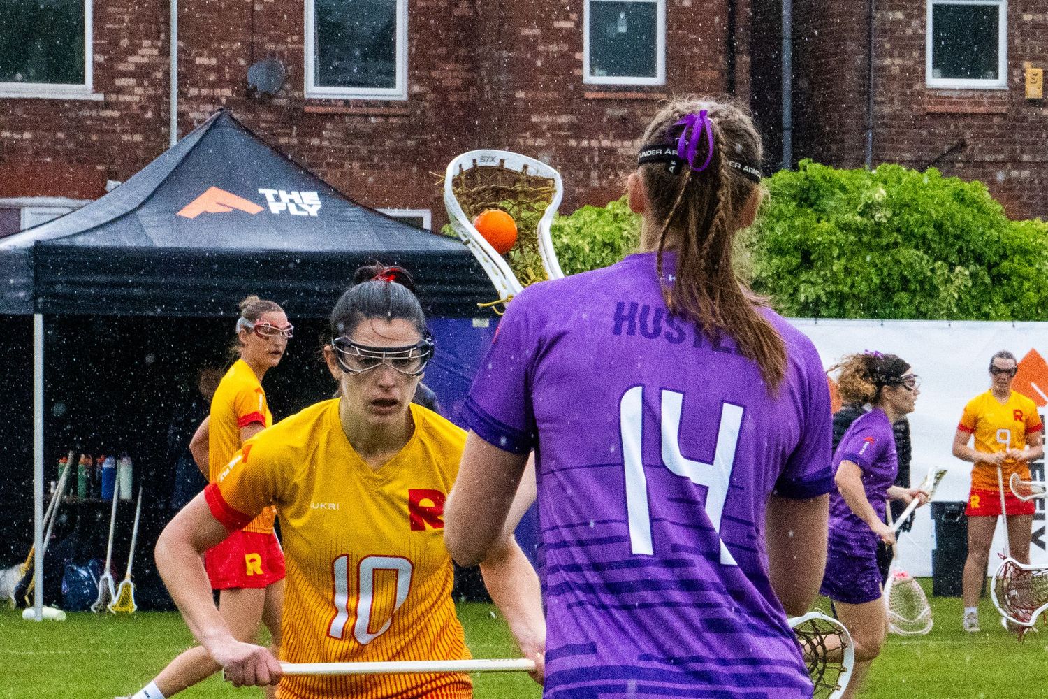 Female lacrosse players face off during outdoor match, one in orange uniform, the other in a purple uniform.