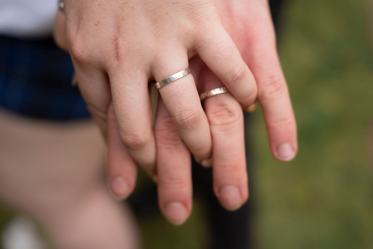 Two hands with matching silver wedding bands touching fingers outdoors on a blurred green background.