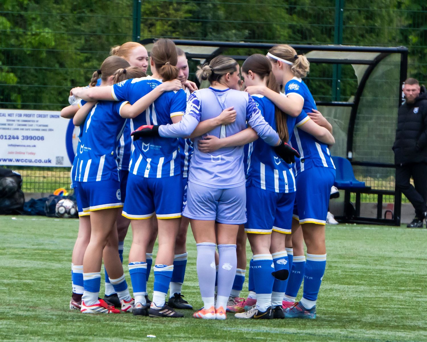 A women's soccer team huddles together in blue and white uniforms on a green field.