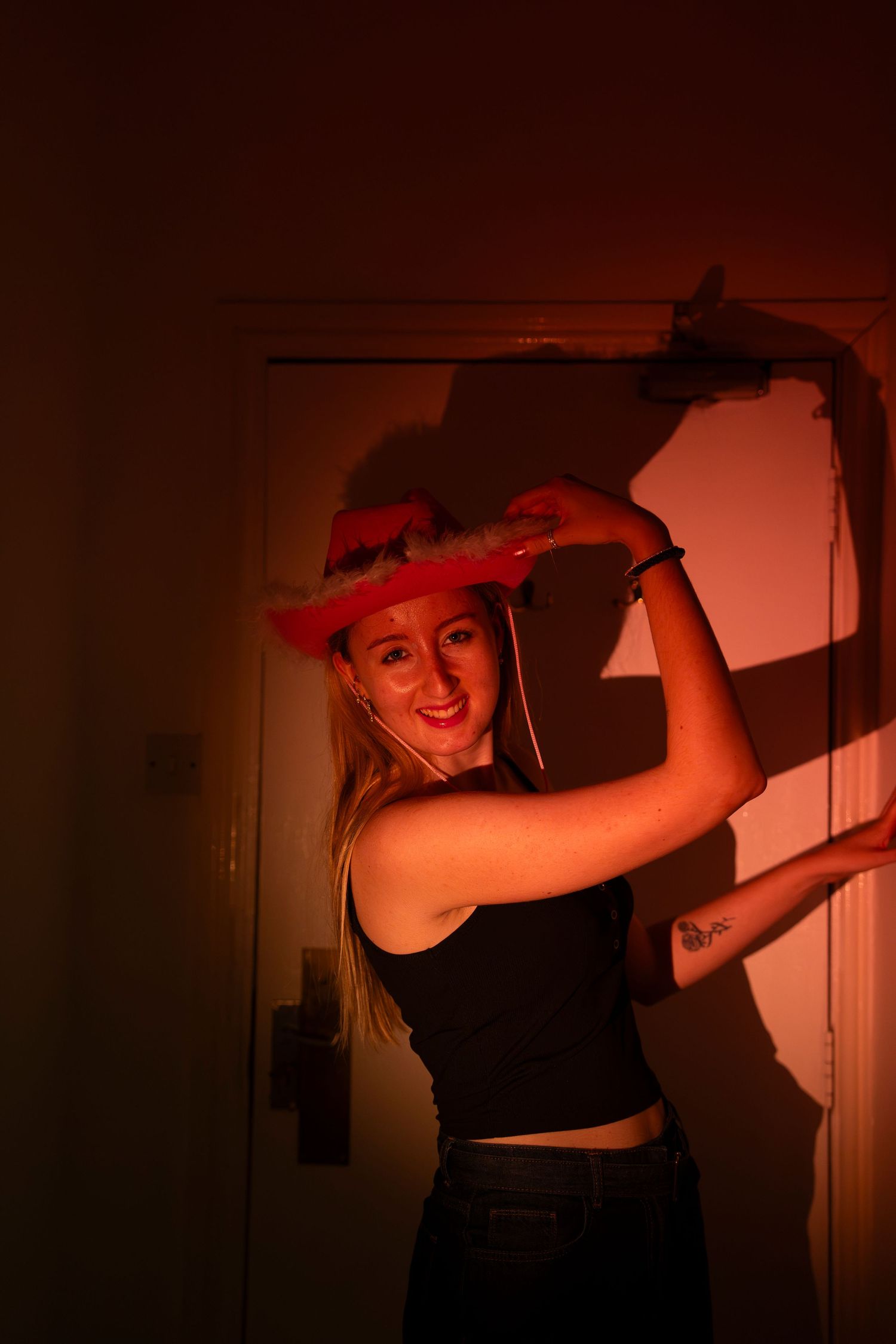 Young blonde woman in black tank top poses playfully wearing a pink cowboy hat under warm red ambient lighting indoors.