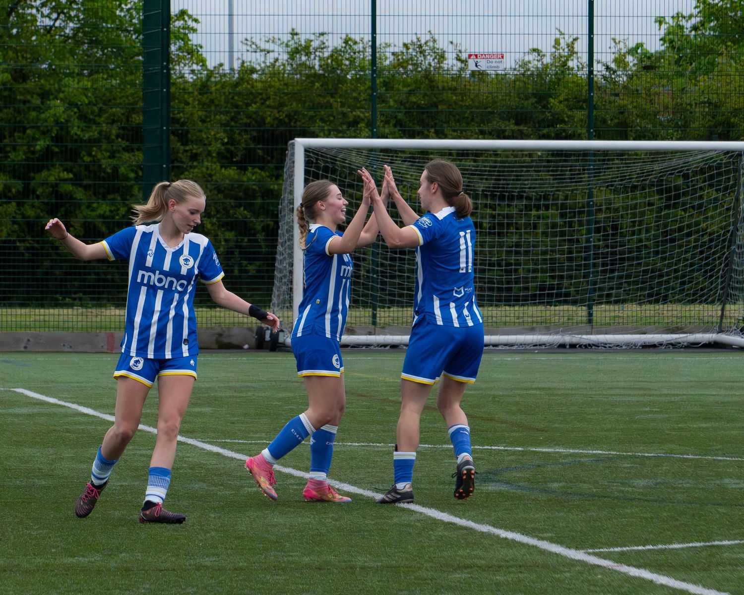Three female football players in blue and white uniforms celebrating together with high fives on a pitch.