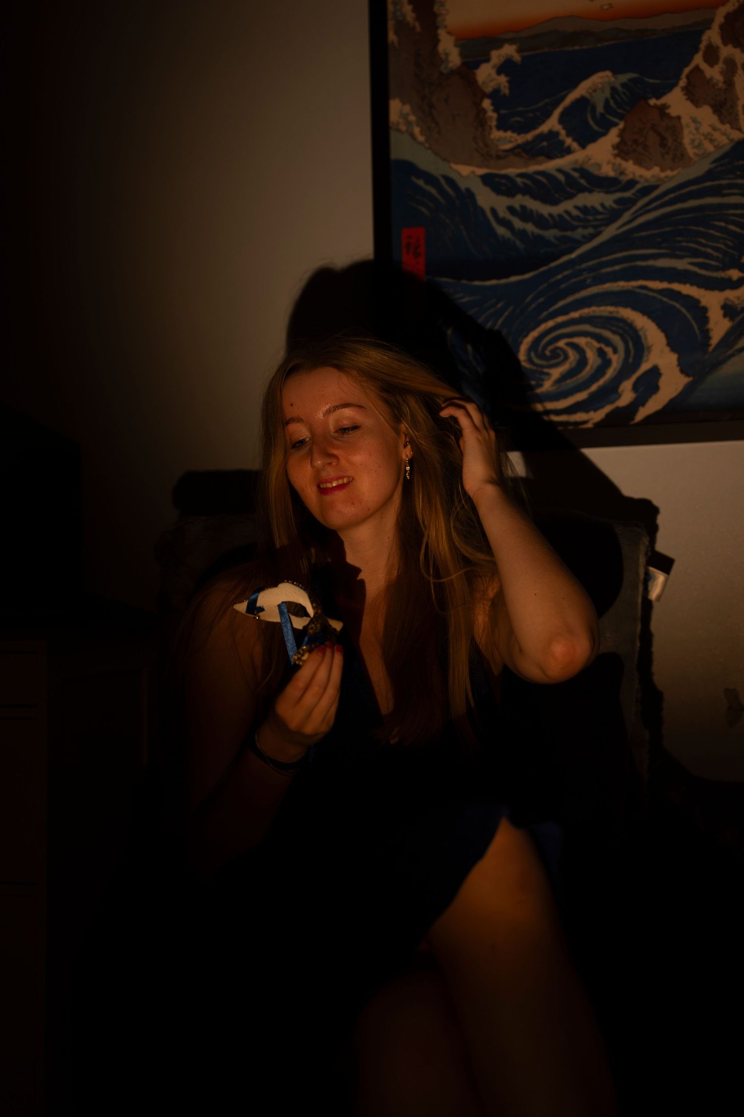 Blonde woman examines small toy figurine in warm candlelit room with Japanese wave wall art.