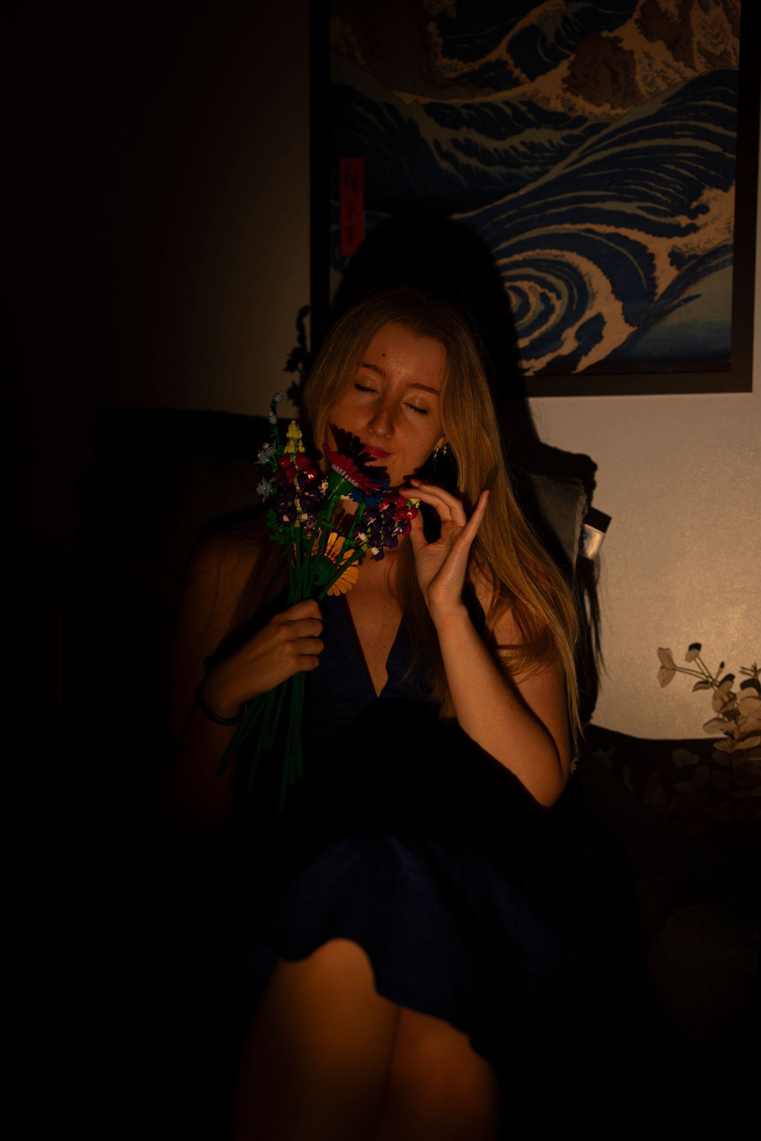 A woman in a dark blue dress holds colorful flowers close to her face in a dimly lit room with wave art.