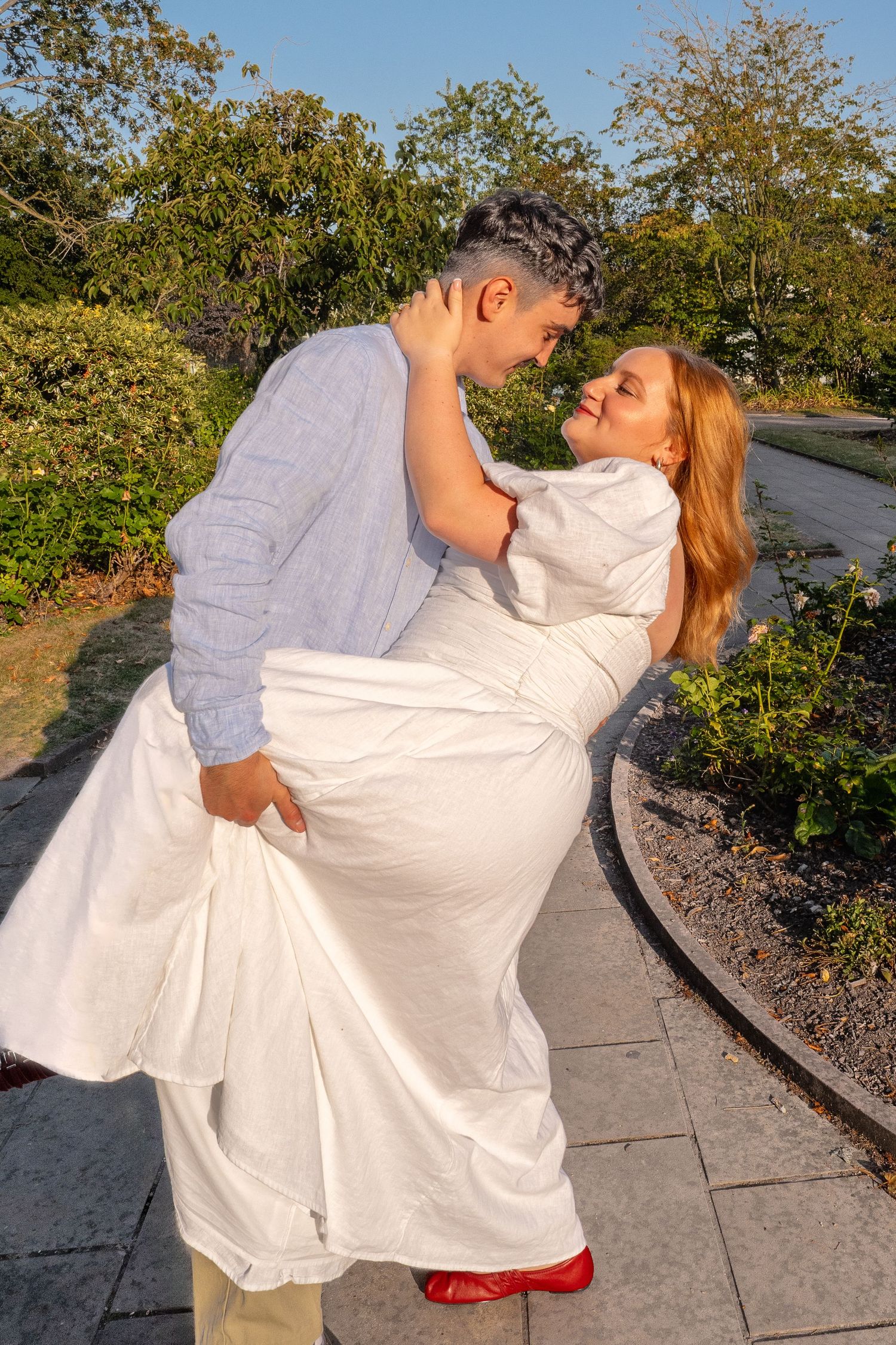 Couple sharing a romantic dip pose in a garden, woman in white dress and red shoes, man in light blue shirt.