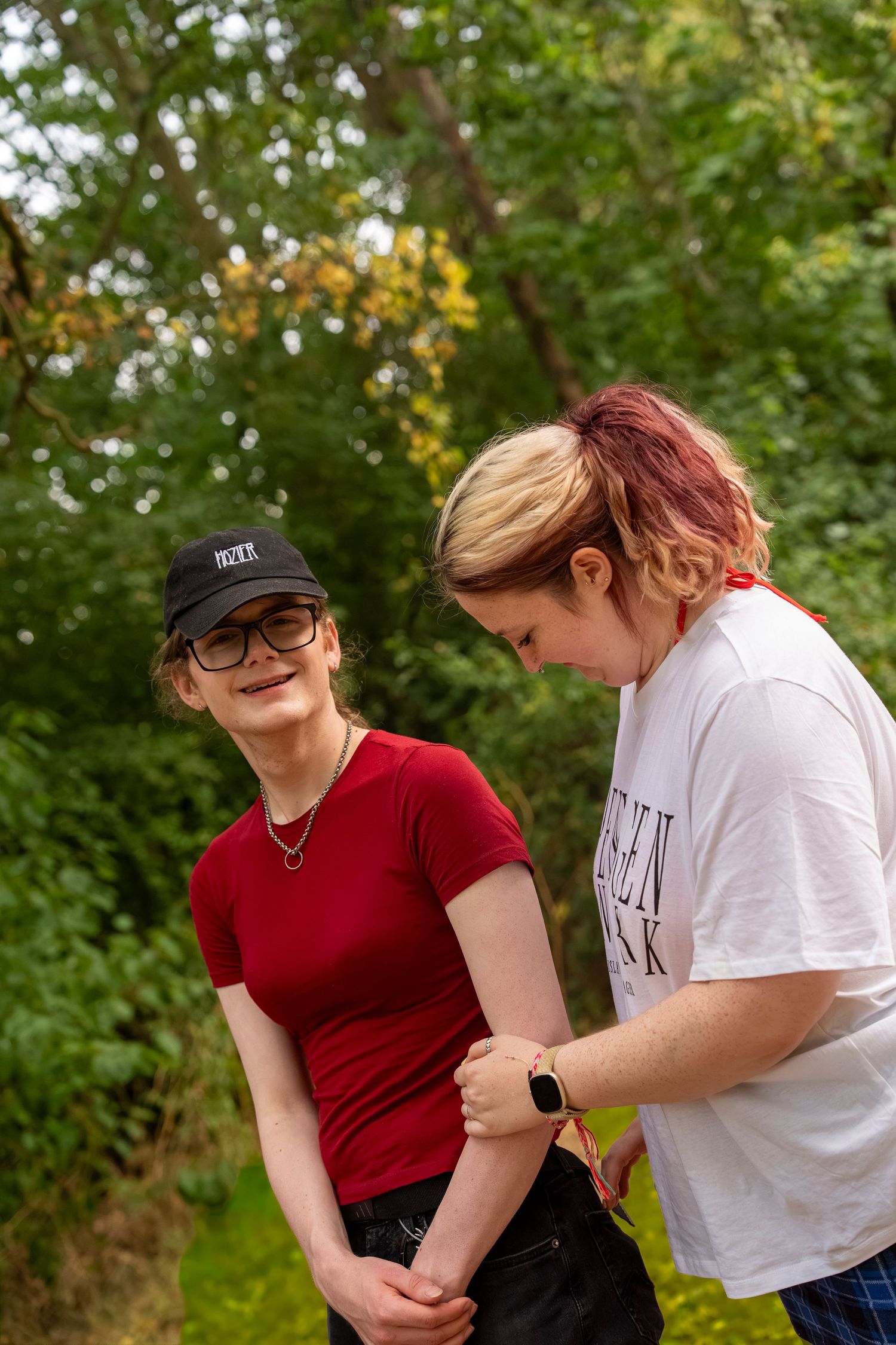Two young women standing outdoors near green trees, one in a red shirt and cap, the other in a white shirt with pink hair.
