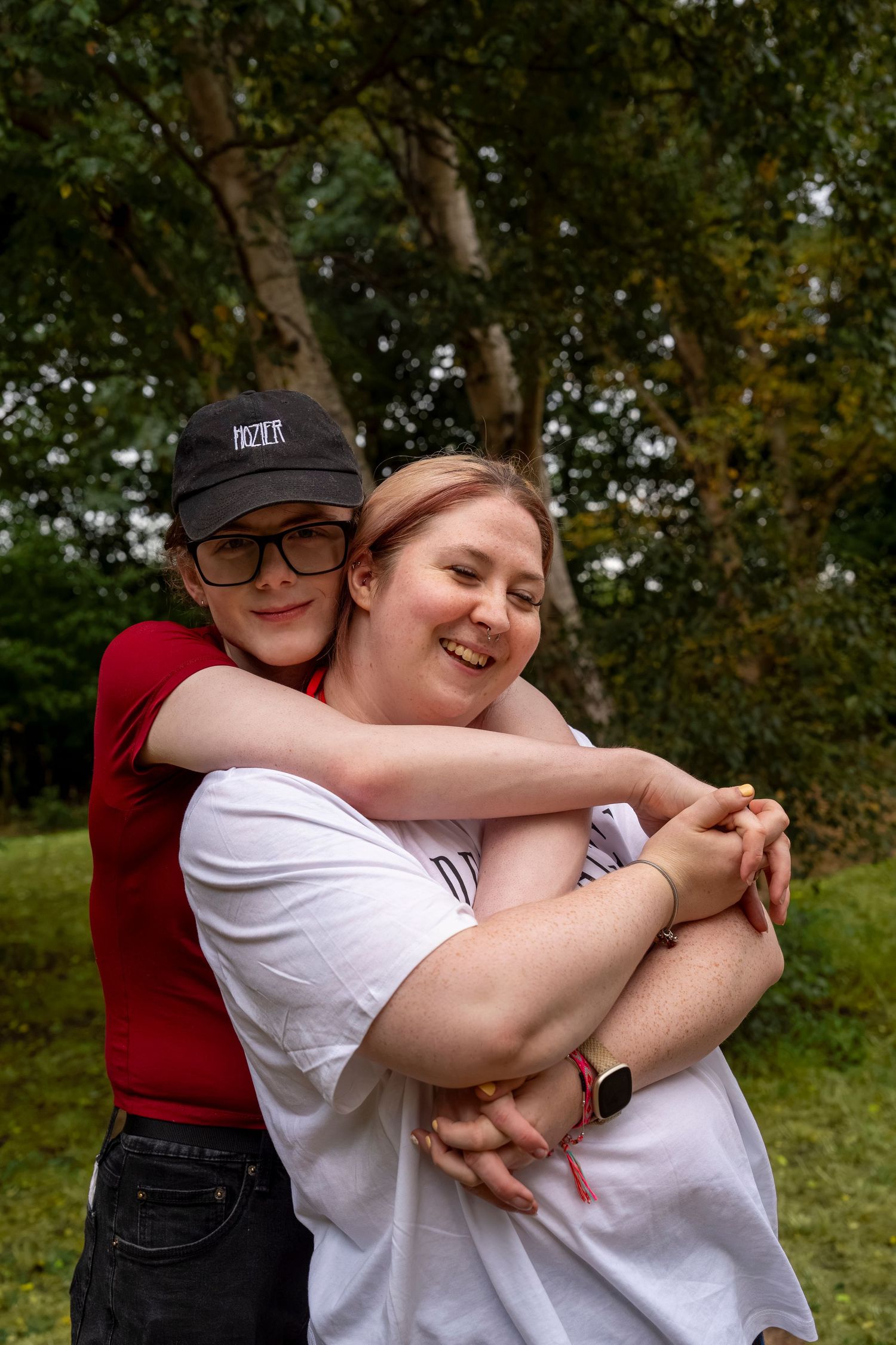 Two young people hugging and smiling outdoors, surrounded by green trees in a park setting.