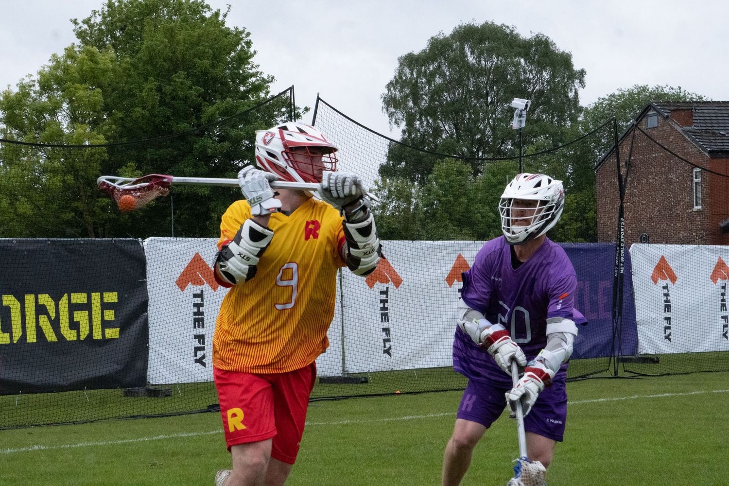 Two male lacrosse players compete during outdoor match in red and purple uniforms.