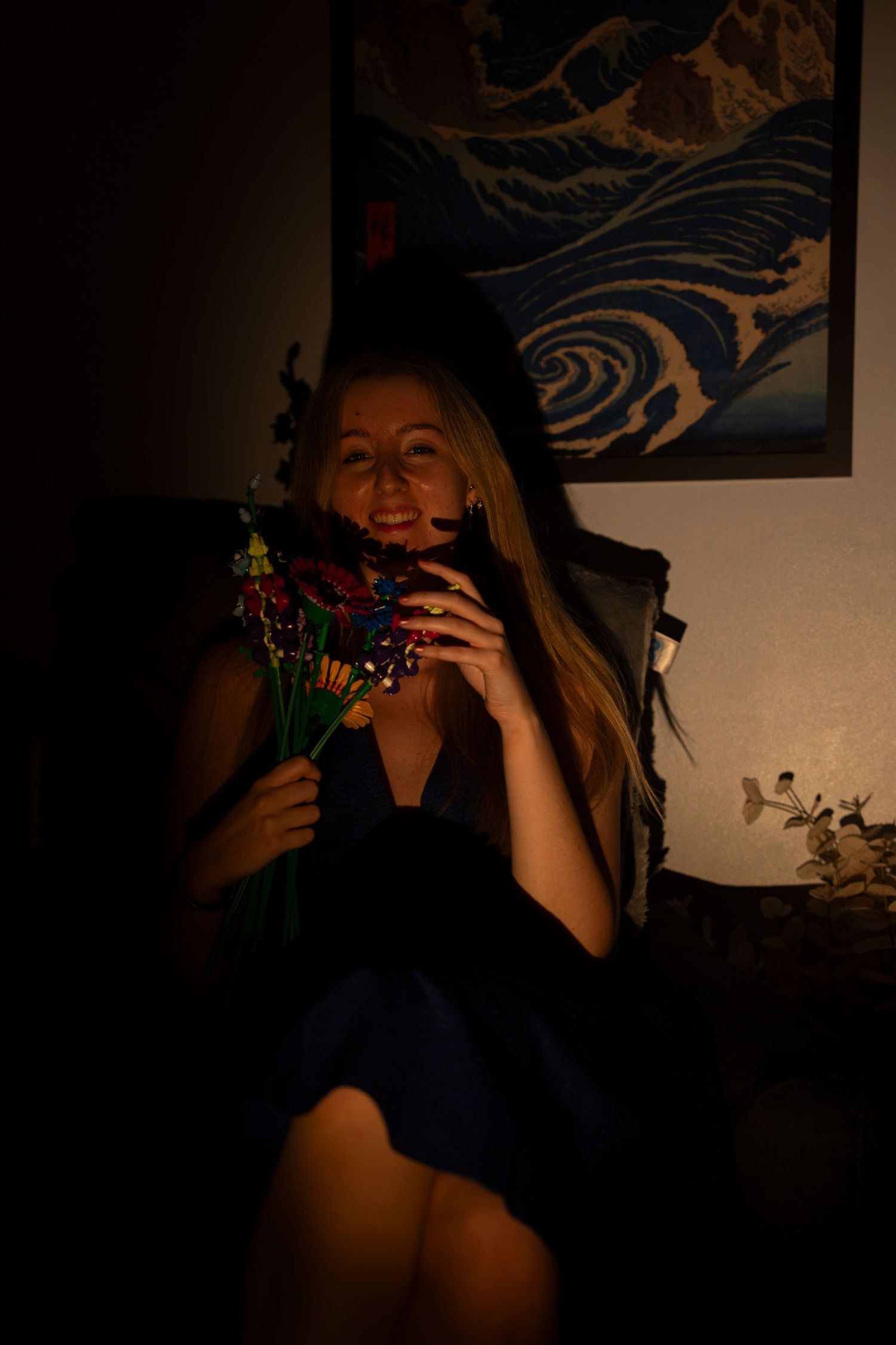 A smiling blonde woman in a dark blue dress holds a colorful bouquet of flowers in a dimly lit room with wave art.