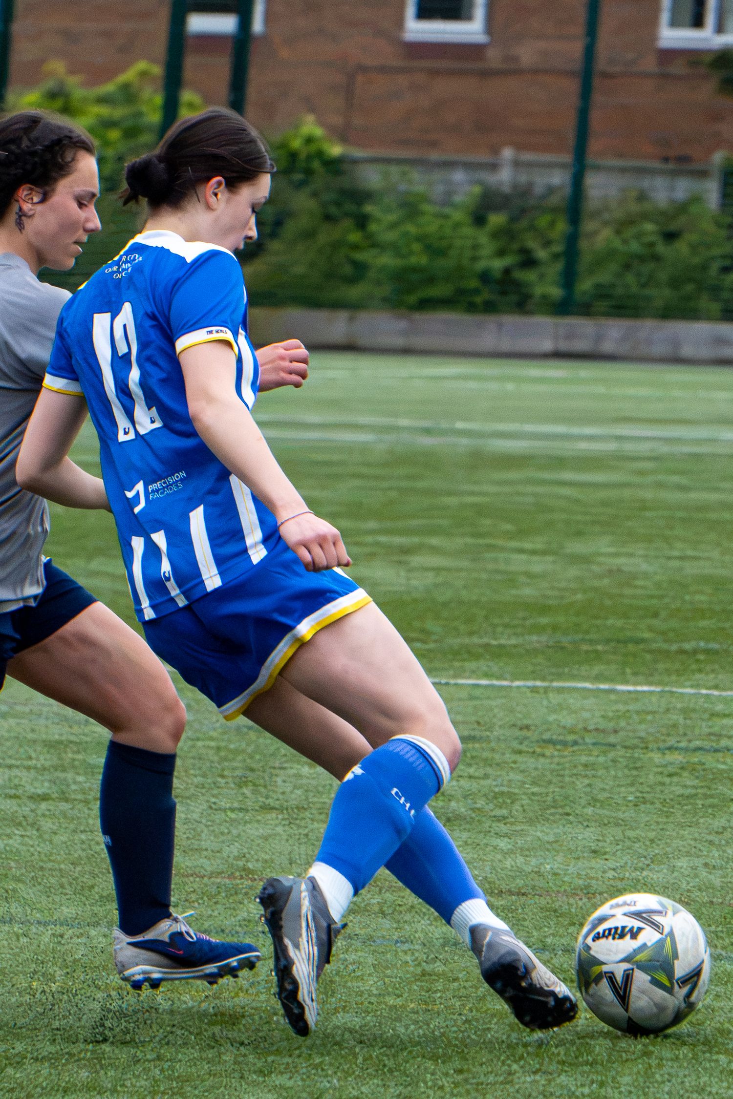 Female soccer player number 12 in blue kit kicking a ball during a competitive women's football match.