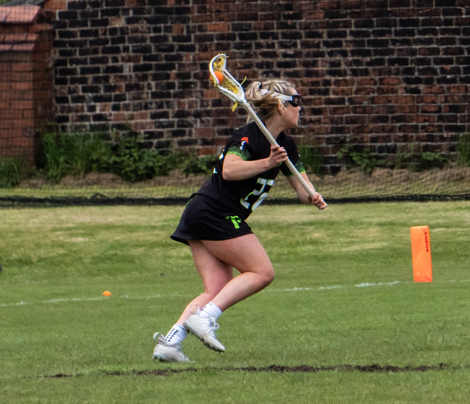 Female lacrosse player in black uniform runs with stick during outdoor game.