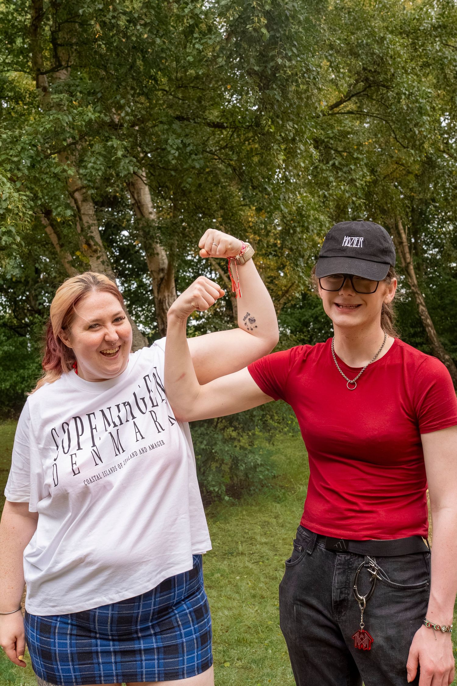 Two women laughing outdoors, one in a Copenhagen Denmark tee and plaid skirt, the other in red flexing her arm tattoo.