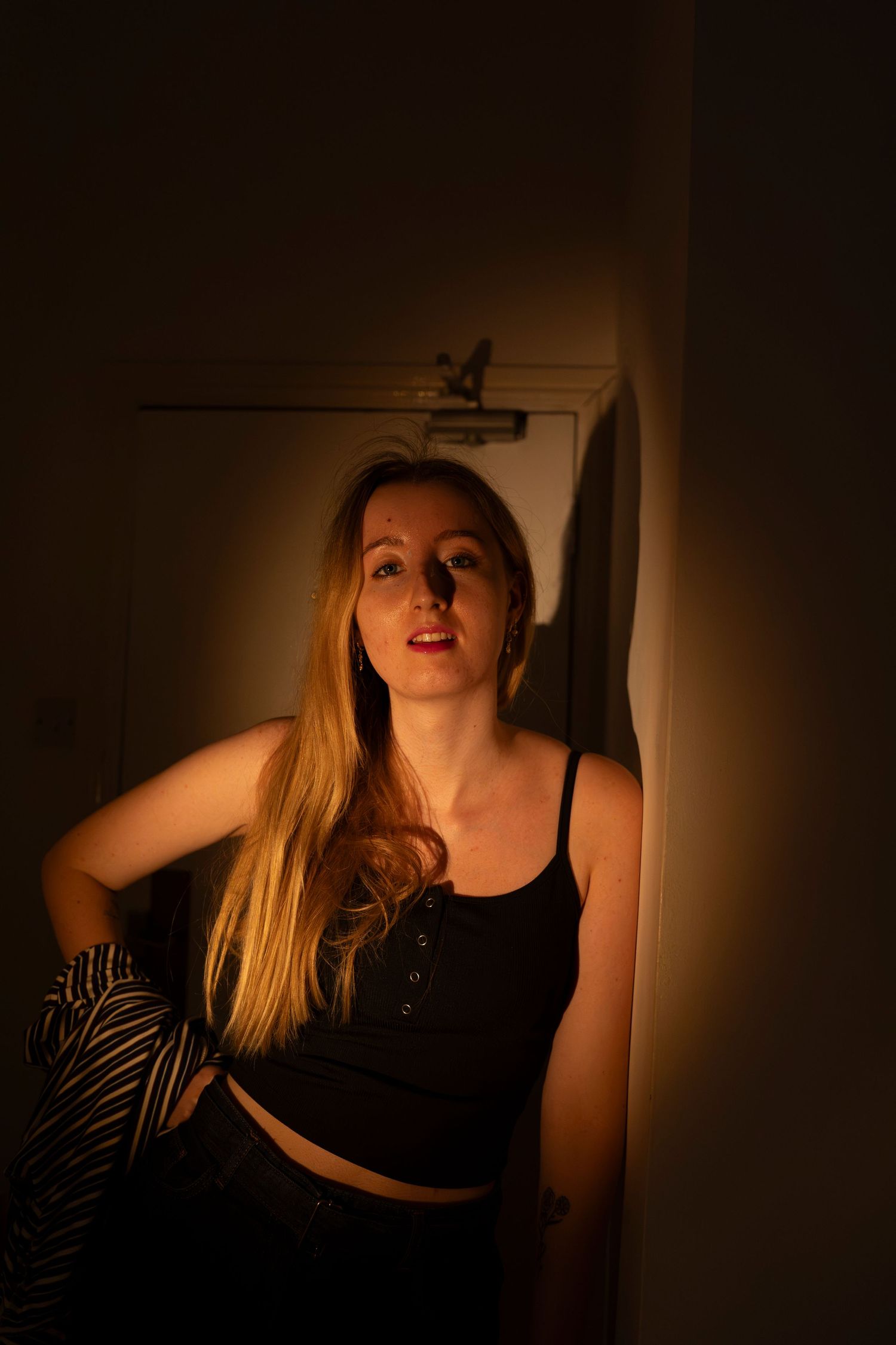 Young blonde woman in black crop top poses against a warmly lit wall in moody amber lighting.