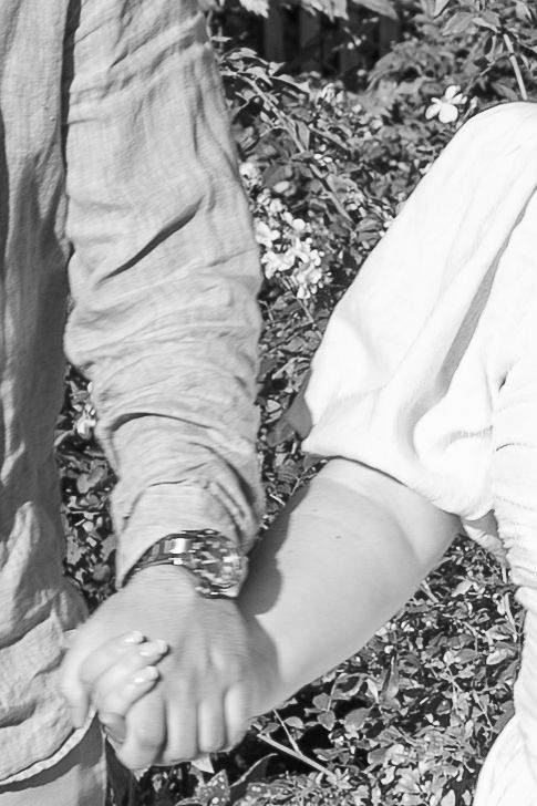 Black and white photo of a couple holding hands outdoors, man wearing a watch, surrounded by flowering plants.