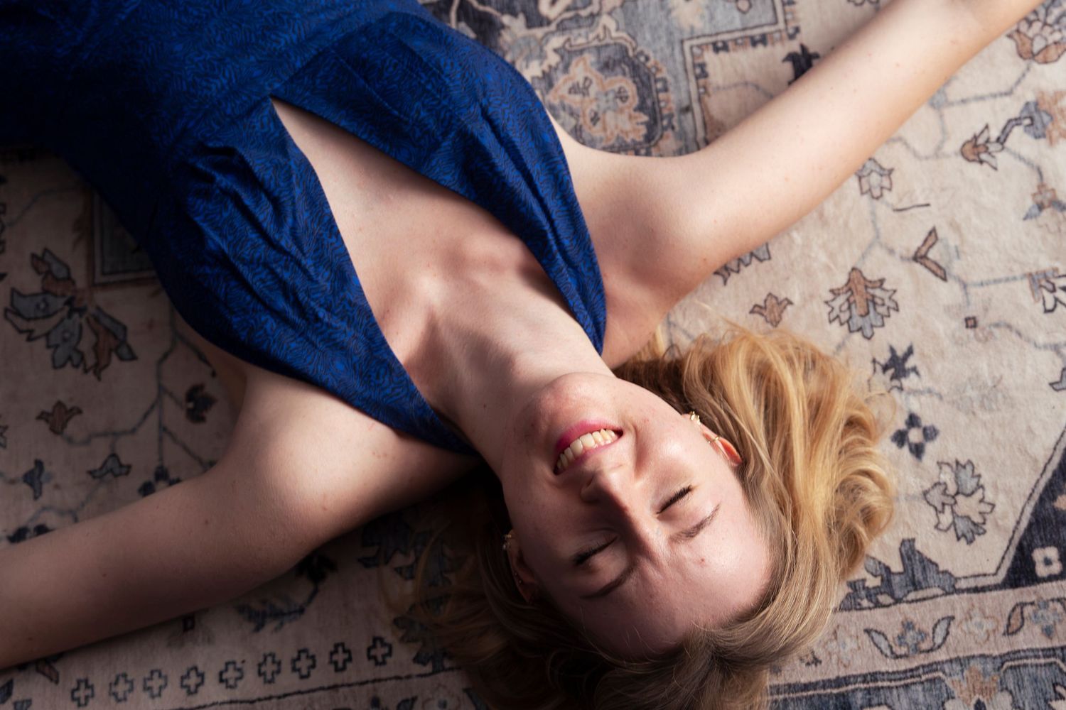 A smiling blonde woman in a blue dress lying on a decorative patterned rug, looking upward with arms outstretched.