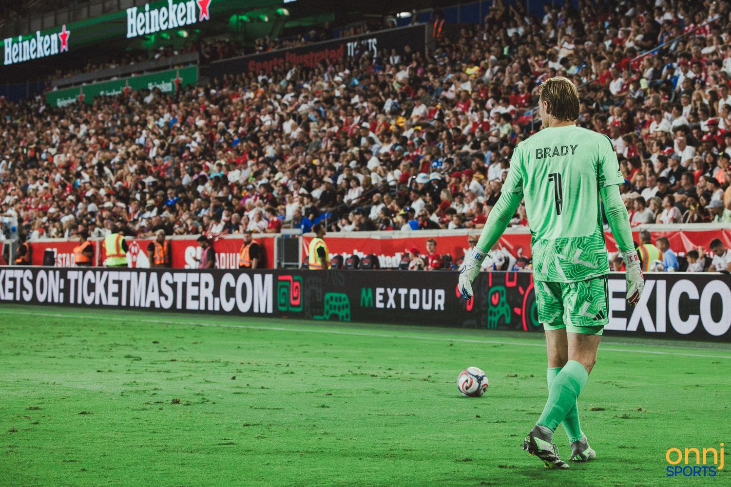 Soccer goalkeeper Brady stands on the field during a packed stadium match with Heineken and Ticketmaster ads visible.