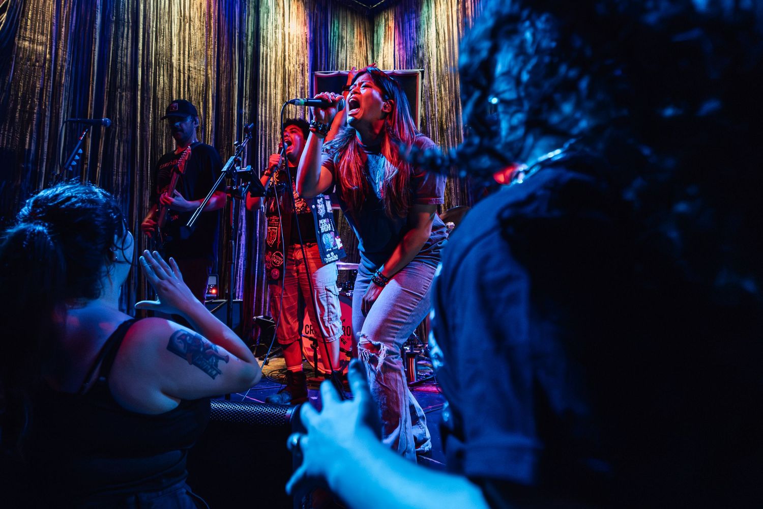 Live band performing on stage with colorful lighting, singer in red outfit, guitarist and bassist visible in vibrant club setting.