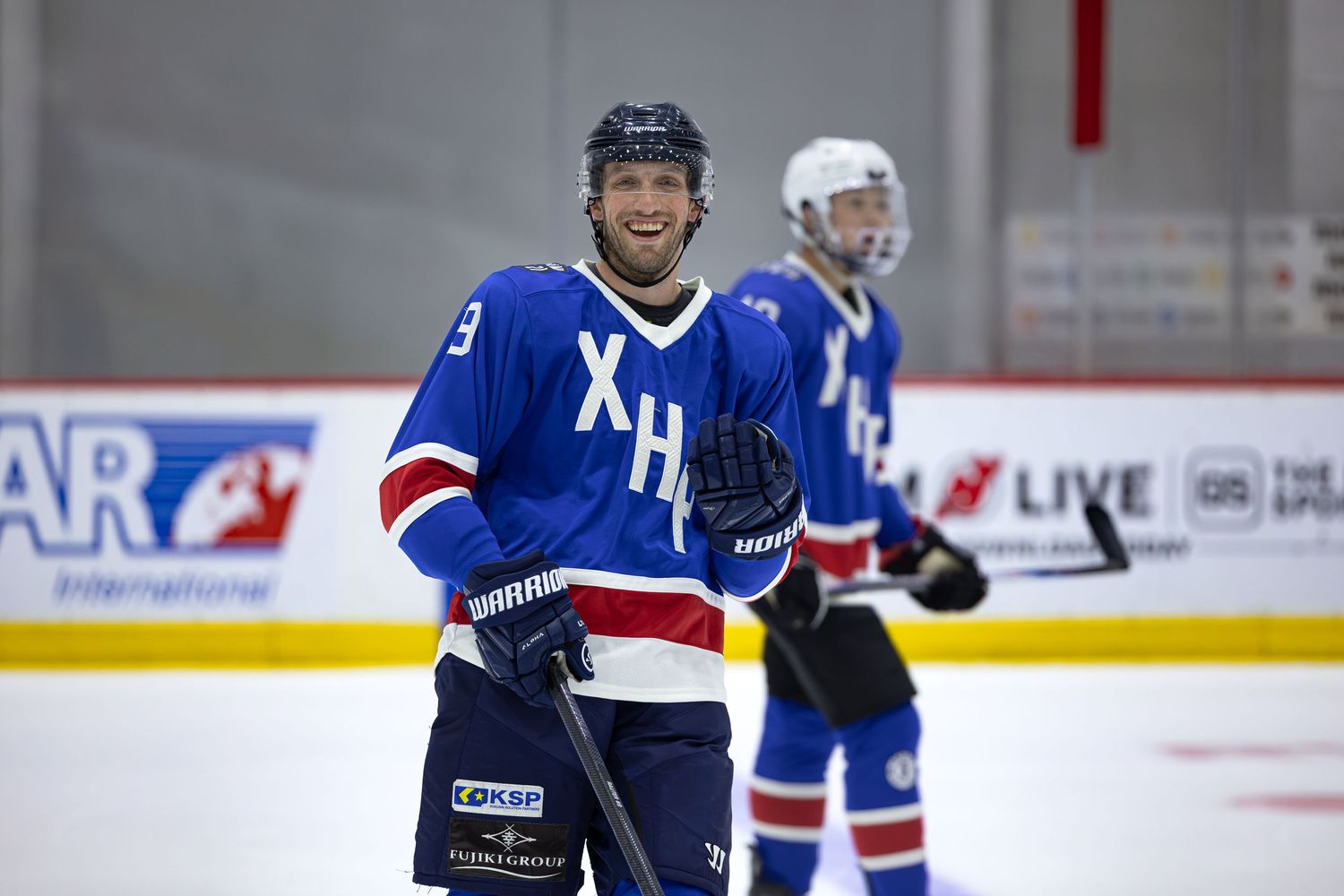 Smiling ice hockey player in blue XH jersey holding a stick on an indoor rink during a game or practice.