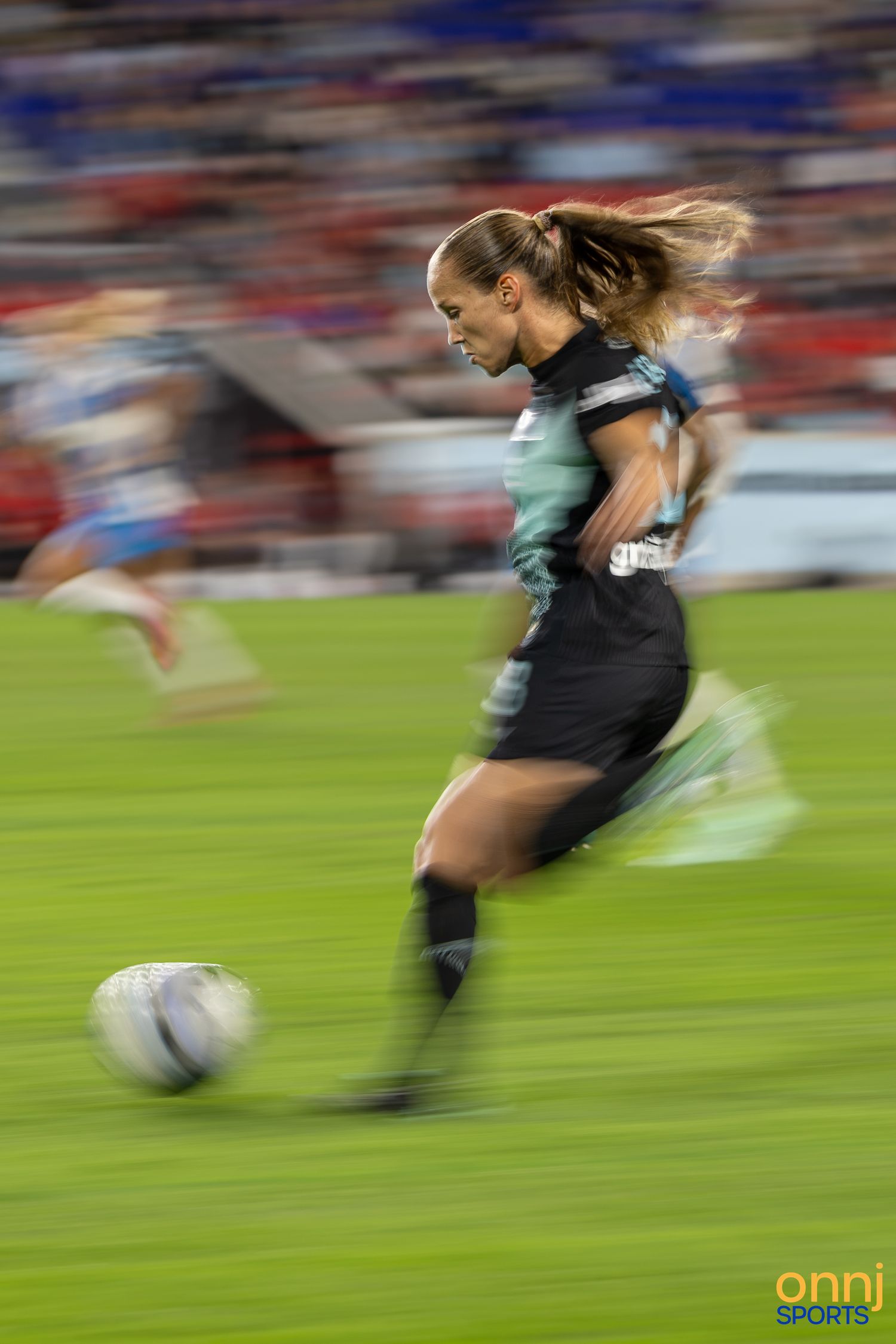 Female soccer player in black uniform sprinting with ball during match, motion blur capturing speed and athleticism.