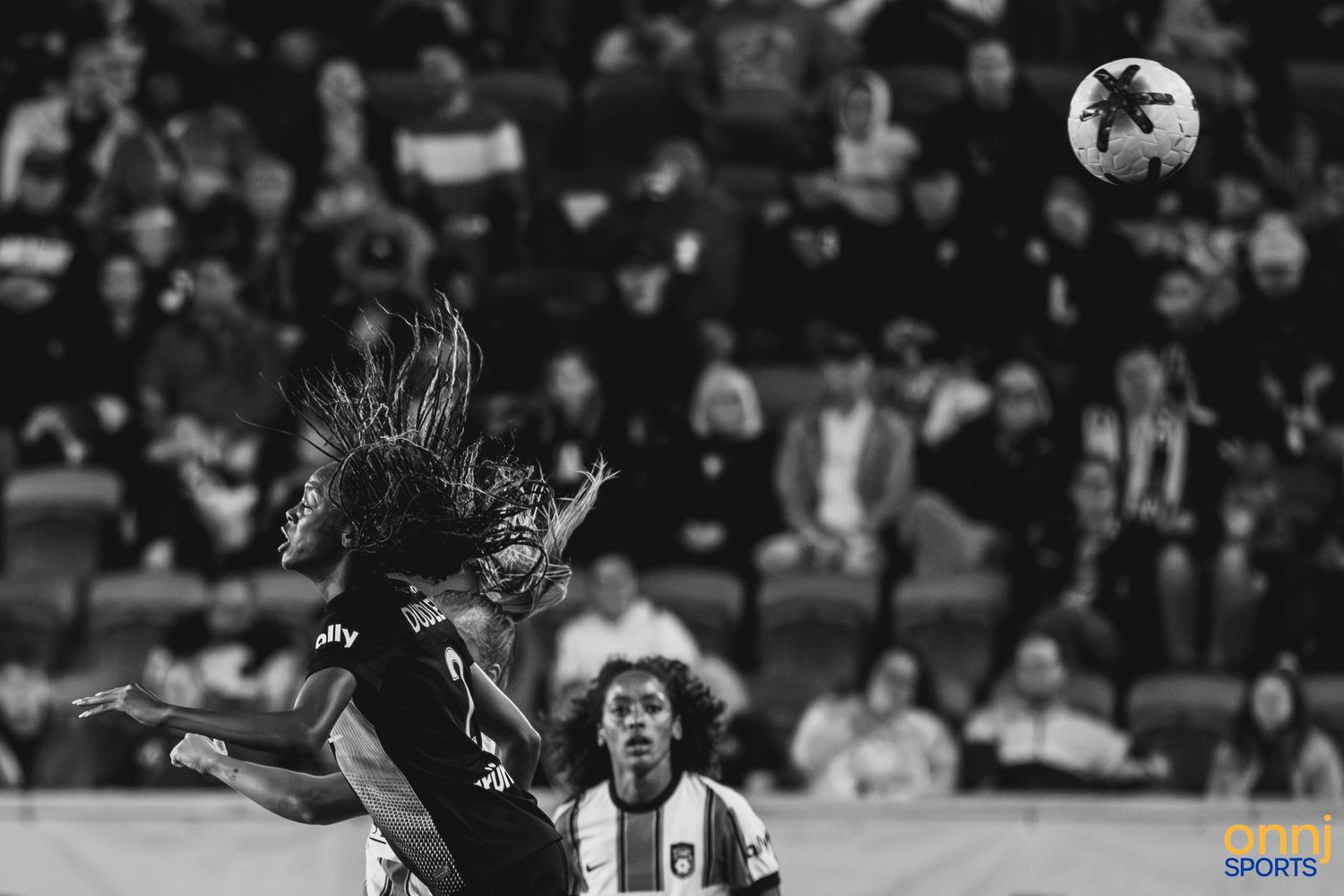 Black and white photo of a soccer player heading the ball during a match with crowd in background.