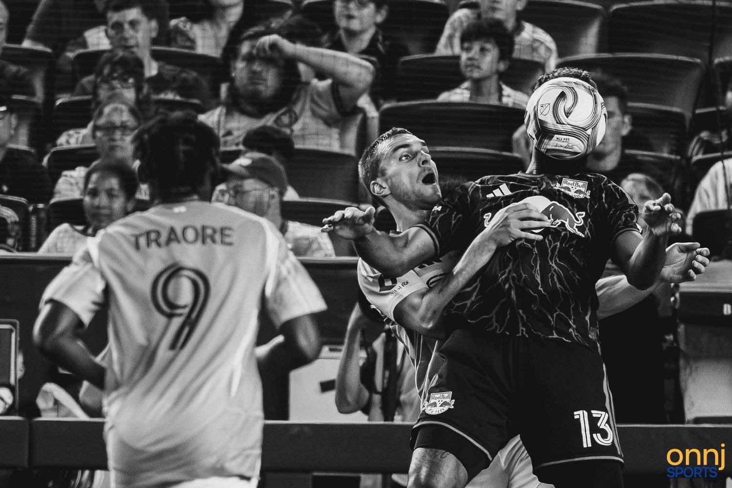 MLS soccer players battle for a header during a match, with player #9 Traore visible in the background.
