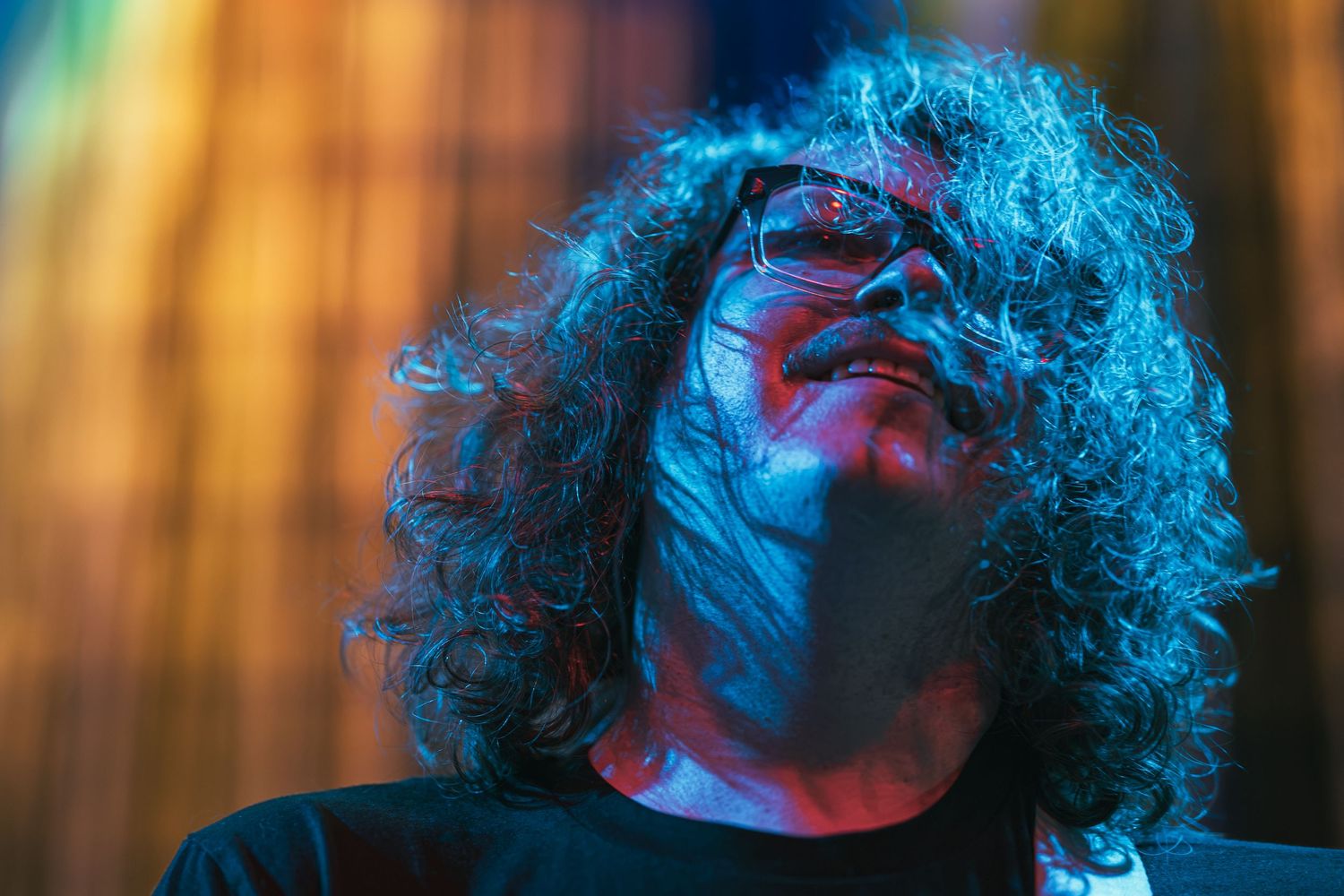 Curly-haired musician with glasses performing on stage under dramatic blue and orange lighting.