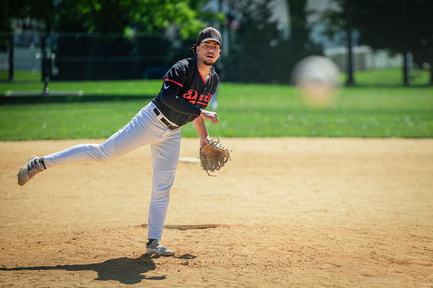 Baseball pitcher mid-throw on a dirt infield, wearing a black uniform with a glove, during an outdoor game.