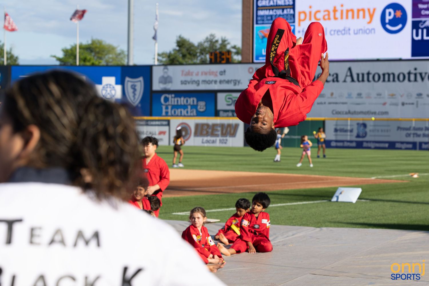 A martial artist in red performs a backflip at a baseball stadium during a Team Black K demonstration event.