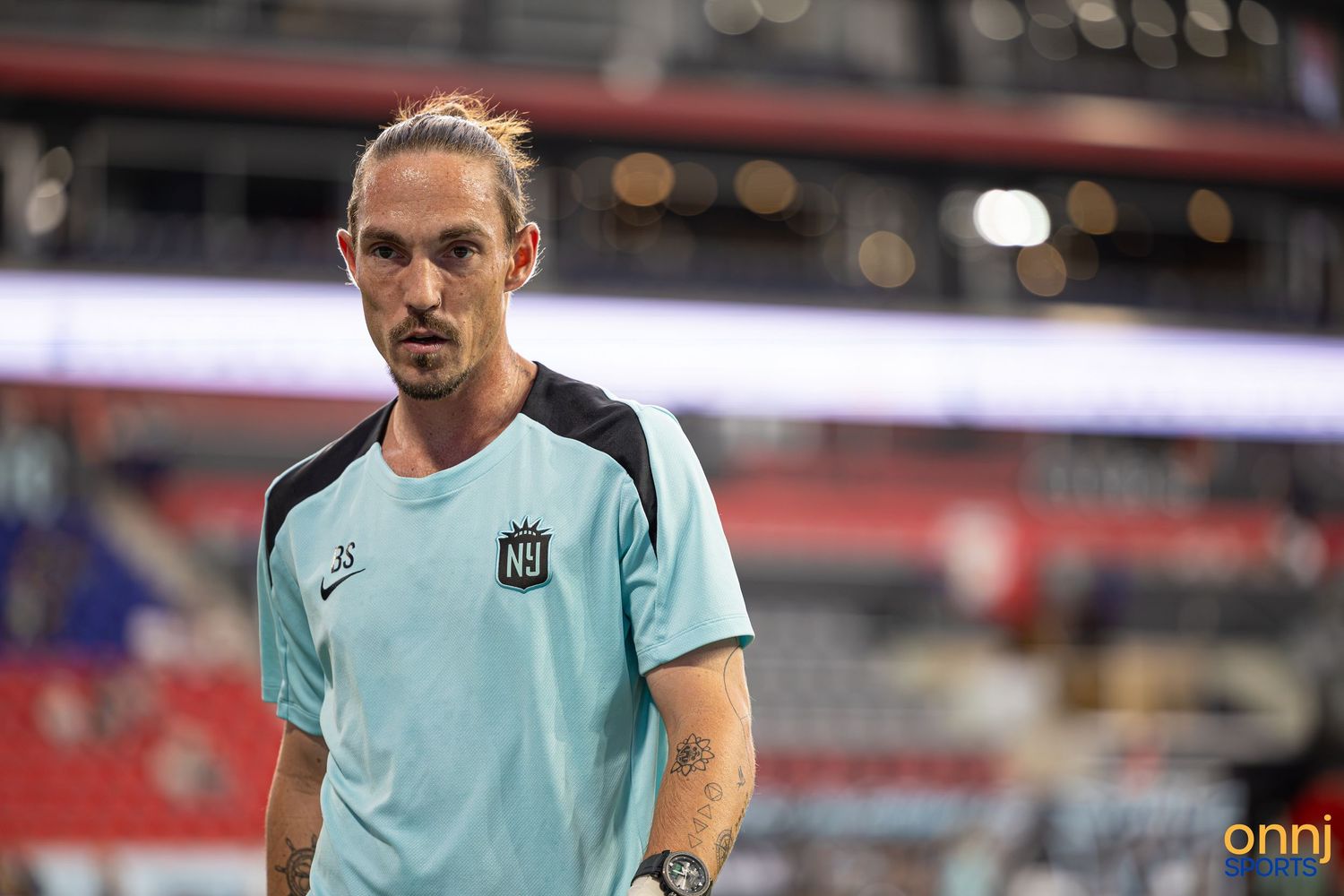 Male soccer player in teal jersey with tattoos stands on stadium field during warm-up session.