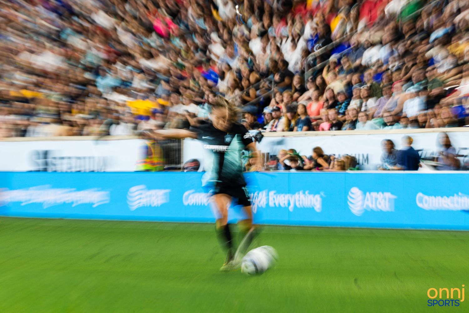 A soccer player kicks a ball on the field before a packed stadium crowd, captured with a dynamic motion blur effect.