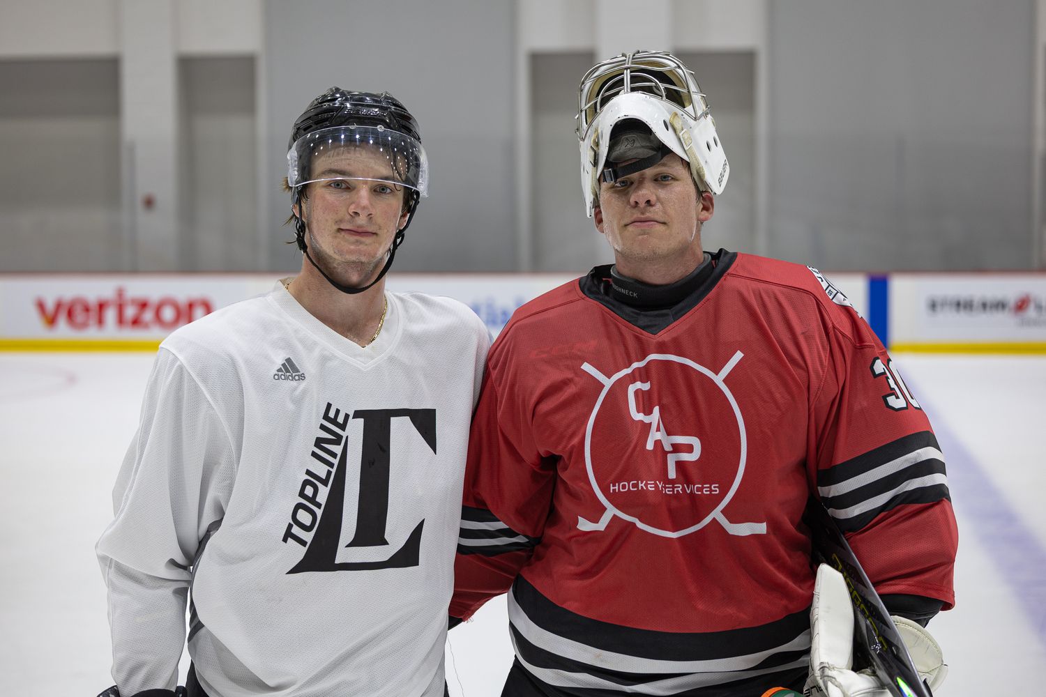 Two hockey players pose together on an ice rink, one in white Topline jersey and one in red goalie gear.