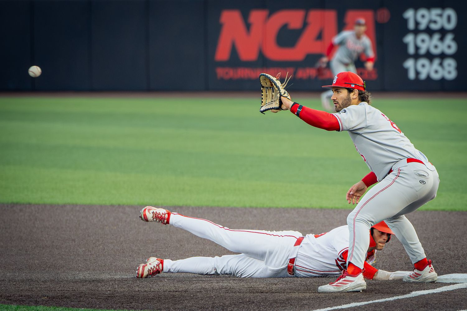 NCAA baseball players in red uniforms during a close play at base, with one player sliding and another reaching for the ball.