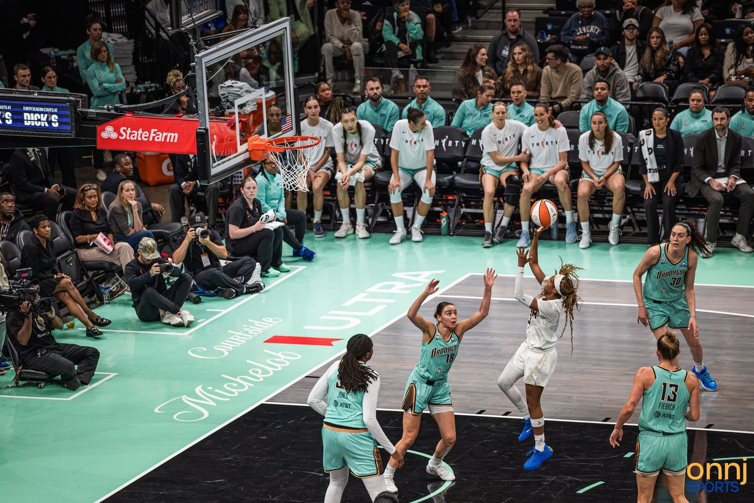 WNBA game action shot of a player driving to the basket against defenders in teal uniforms at an indoor arena.