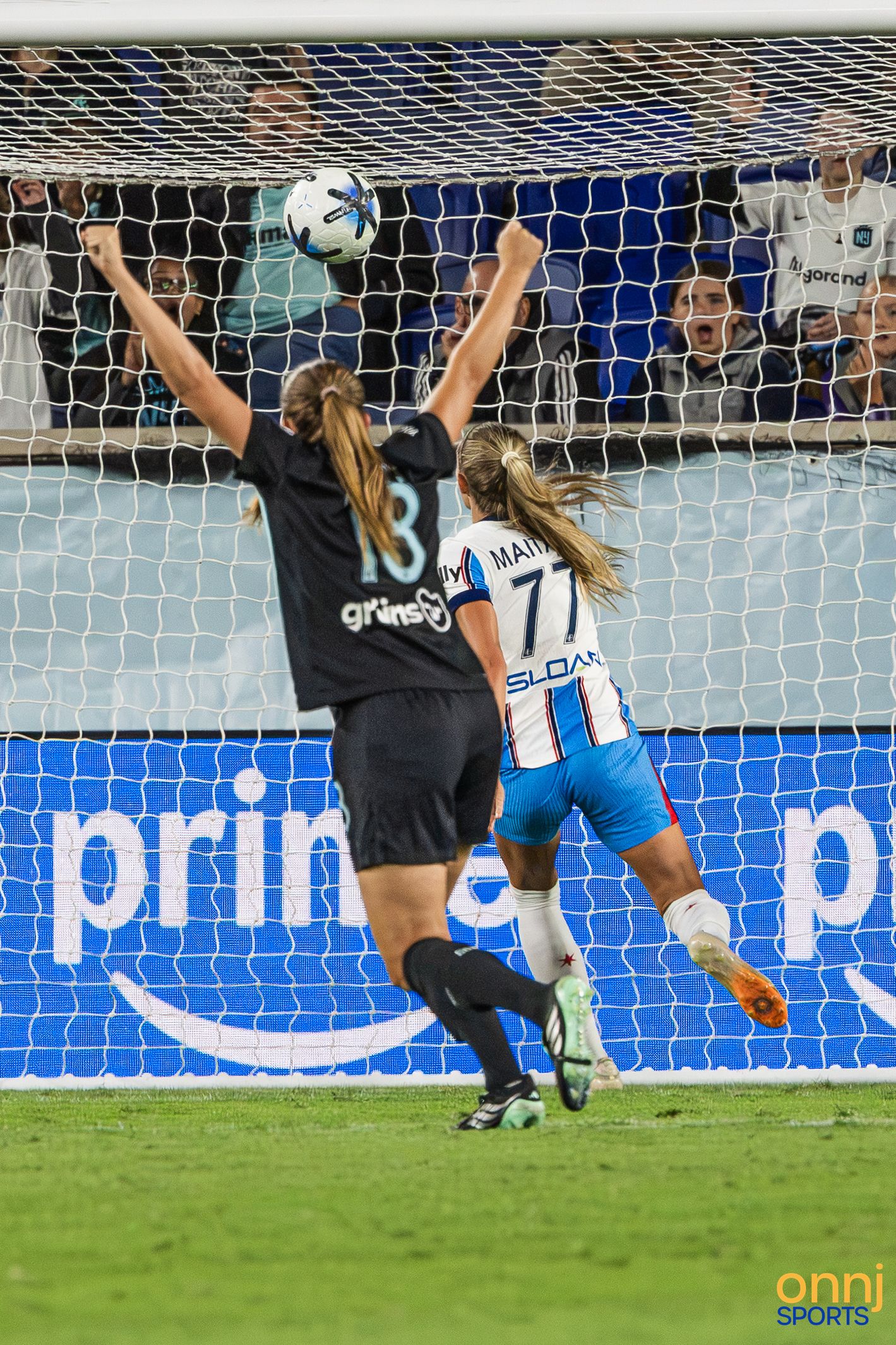 Female soccer goalkeeper in black jersey #13 leaps to save a shot on goal during a night match.