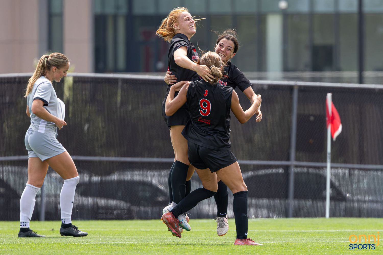 Female soccer players in black uniforms celebrate a goal with a joyful group hug on a green field.