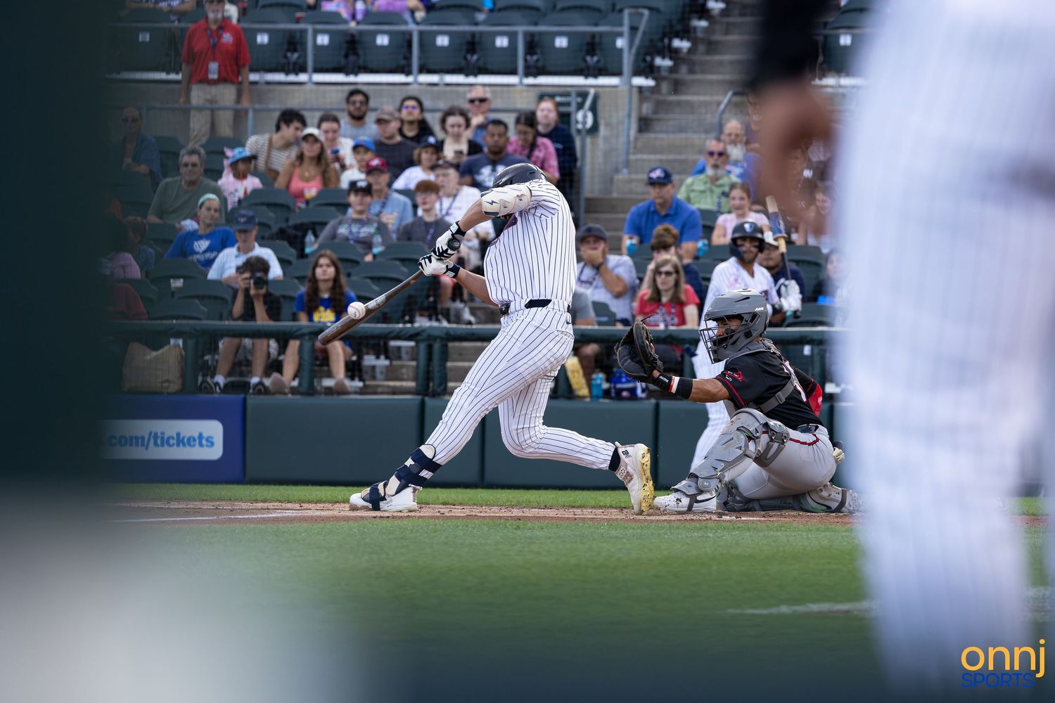 Baseball batter swings at pitch during minor league game with catcher and crowd in background.