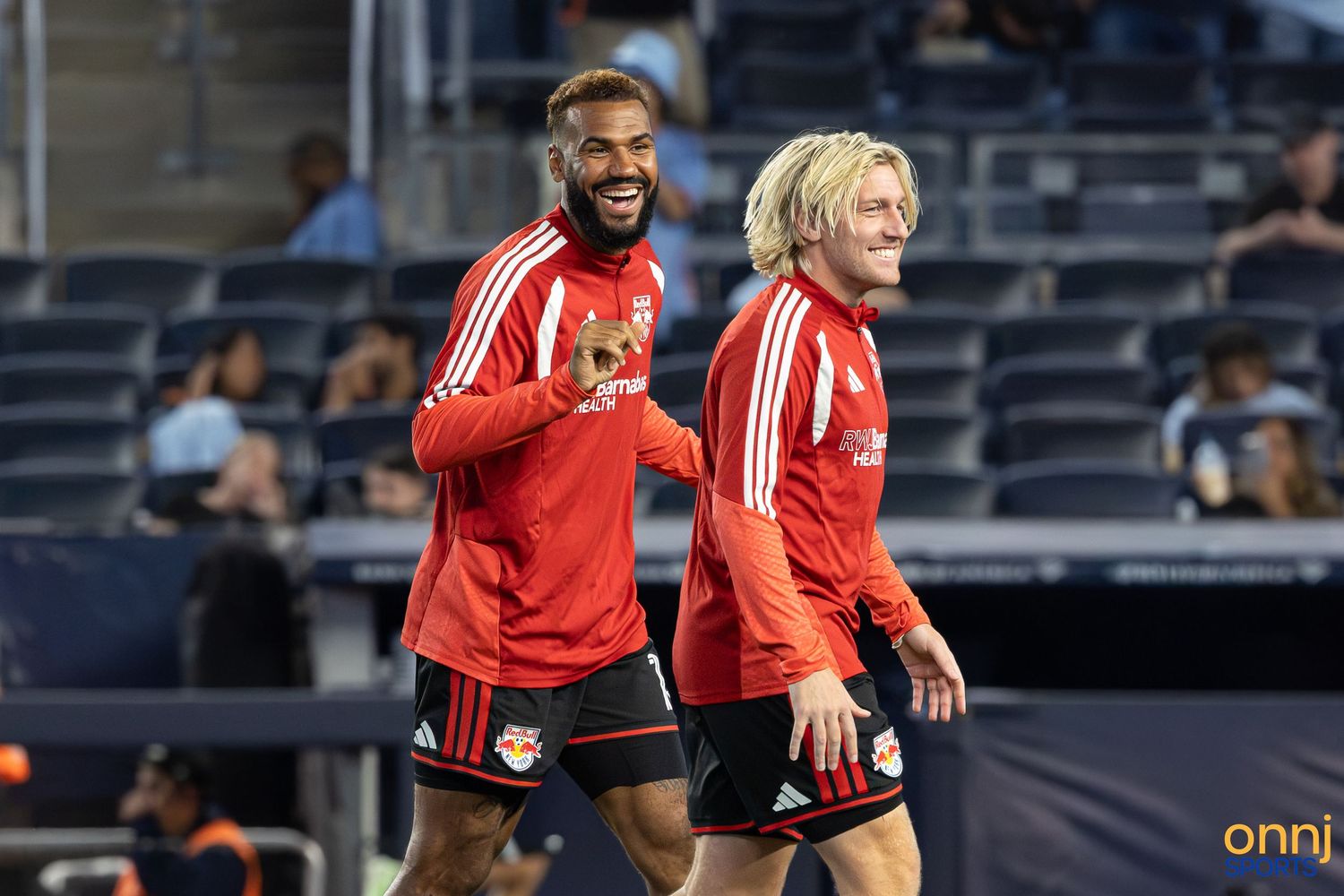 Two Red Bulls soccer players smiling and laughing during warm-ups on the field at night.